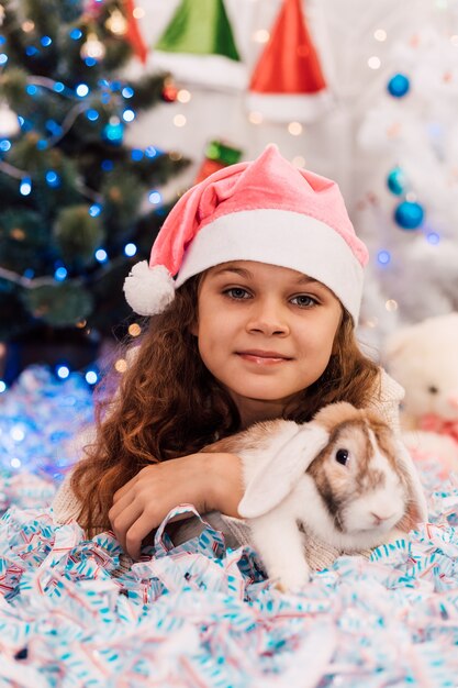 Año nuevo, una niña con un gorro de Papá Noel rosa se encuentra cerca de un árbol de Navidad y acaricia un conejo