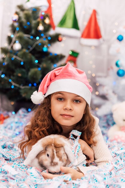 Año nuevo, una niña con un gorro de Papá Noel rosa se encuentra cerca de un árbol de Navidad y acaricia un conejo