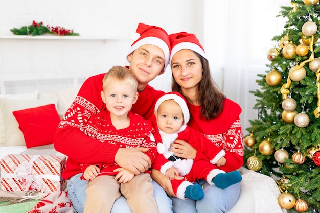 Año nuevo o Navidad, una familia feliz con dos niños en la cama en casa junto al árbol de Navidad en suéteres rojos sonriendo, abrazándose y besándose felicitando por las vacaciones