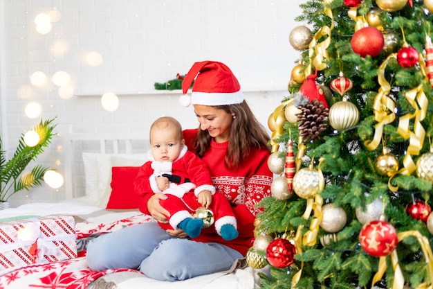 Año nuevo o Navidad, una joven madre con un bebé en la cama en casa junto al árbol de Navidad con un disfraz de Santa Claus abrazándose y esperando las vacaciones sonriendo