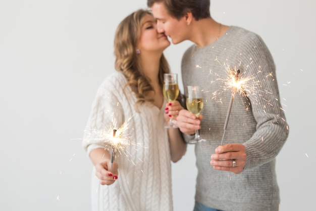 Año nuevo, vacaciones, concepto de fecha - Pareja amorosa sosteniendo luces de bengala y copas de champán sobre pared blanca, primer plano