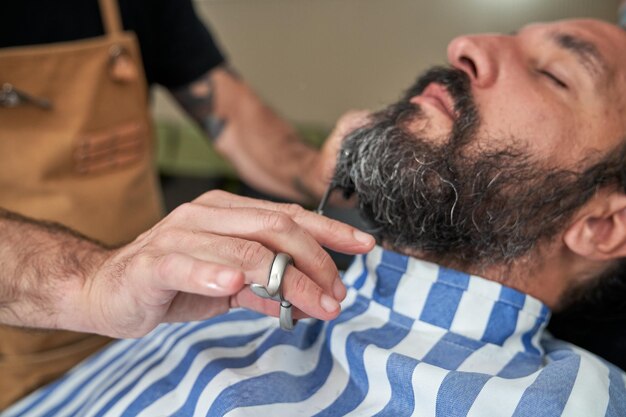 Anonymer Friseur, der während der Arbeit im Friseursalon mit einer Schere die Haare eines bärtigen männlichen Kunden schneidet