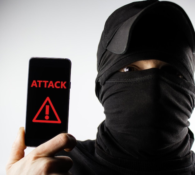 Anonymer Hacker hält ein Smartphone mit der Aufschrift ATTACK auf dem Bildschirm Das Konzept des TelefonbetrugsExtortionMalwareCybercrime