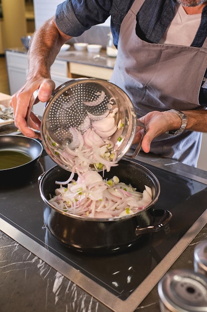 Foto anonymer männlicher koch gießt gewaschene, gehackte zwiebeln aus dem sieb in die bratpfanne auf dem herd, während er in der modernen küche kocht