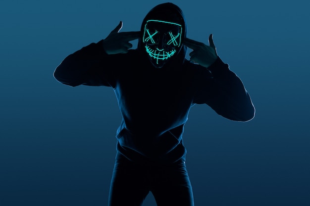 Anonymer Mann im schwarzen Hoodie, der sein Gesicht hinter einer Neonmaske versteckt