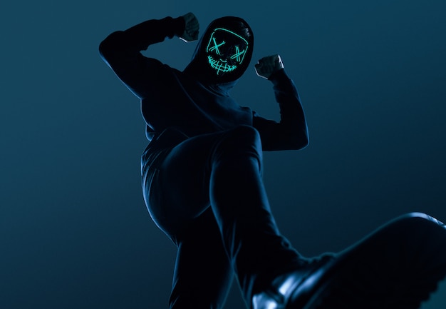 Anonymer Mann im schwarzen Hoodie, der sein Gesicht hinter einer Neonmaske versteckt