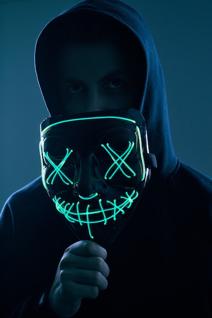 Anonymer Mann im schwarzen Hoodie, der sein Gesicht hinter einer Neonmaske versteckt