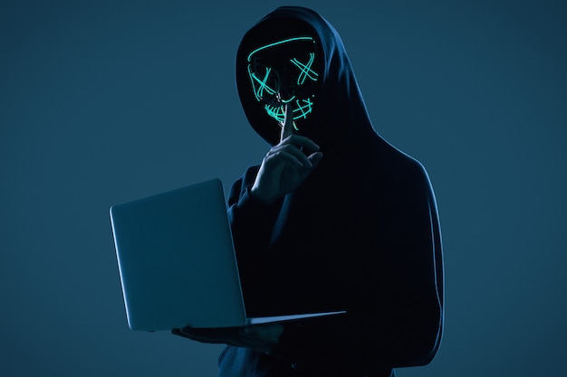 Foto anonymer mann in einem schwarzen hoodie und in einer neonmaske, die in einen computer zerhacken