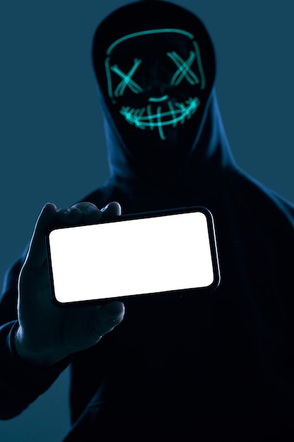 Foto anonymer mann in einem schwarzen hoodie und in einer neonmaske, die in einen smartphone zerhacken