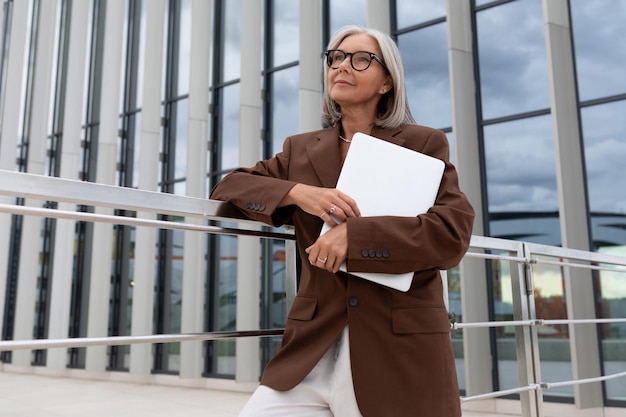 años, exitosa, delgada, mujer de negocios canosa, vestida con estilo, sostiene una computadora portátil mientras