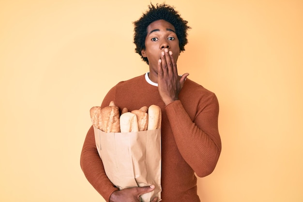 Ansehnlicher afroamerikanischer Mann mit Afrohaaren hält eine Papiertüte mit Brot und bedeckt den Mund mit der Hand schockiert und fürchtet sich vor einem Fehler überrascht Ausdruck