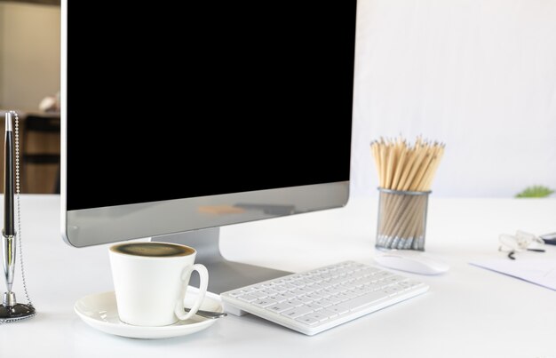 Ansicht des Büroraumcomputerdesktops mit leerem Bildschirm und weißer Tasse schwarzem Kaffee, Stift, Bleistift, Klemmbrett Spott oben. Verwendung als Geschäfts- und Arbeitskonzept.