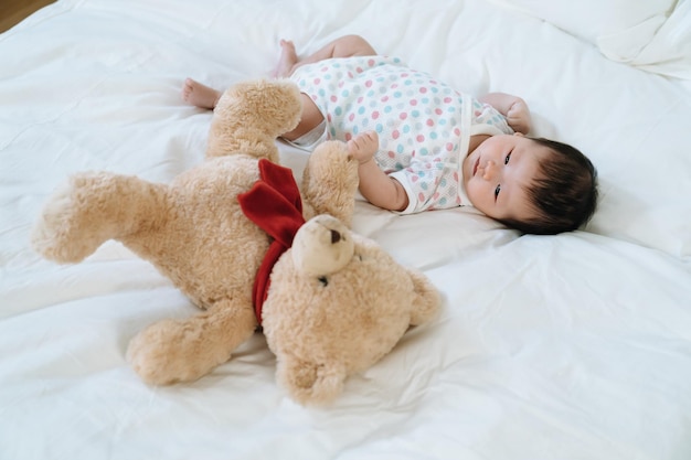 Ansicht des schönen asiatischen Babys, das auf weißer Bettwäsche mit Teddybär liegt. weibliches neugeborenes im bodysuit schaut unschuldig auf stofftier.