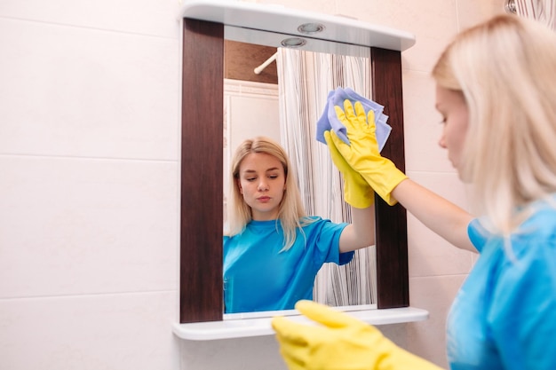 Ansicht eines jungen blonden Mädchens, das gelbe Schutzhandschuhe trägt, die zu Hause mit Spray reinigen und mit einem Rakel den Spiegel im Badezimmer waschen Service- und Reinigungskonzept