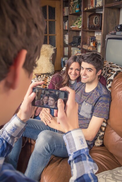 Ansicht eines jungen Freundes, der Fotos mit einem Smartphone zu einem Hipster-Teenager-Paar macht, das auf dem Sofa sitzt