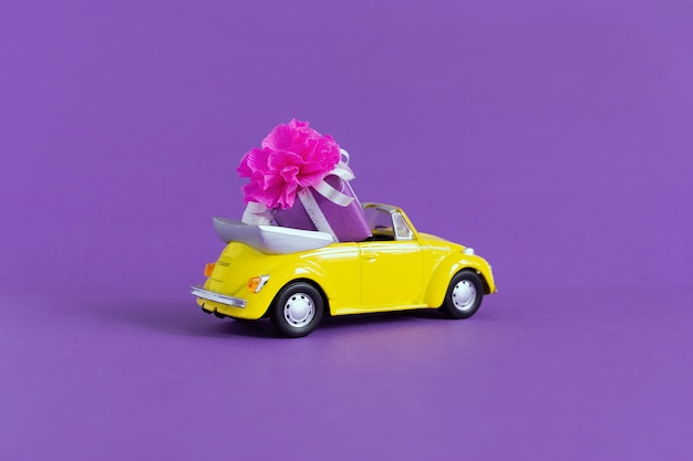 Ansicht eines kleinen bunten gelben Autos, in dem es eine Geschenkbox mit einer Schleife auf einem Purpur gibt. Konzept Urlaub, Transport, Valentinstag, Lieferung