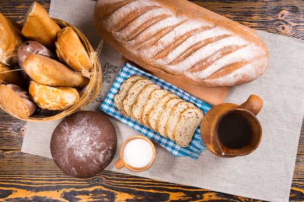 Ansicht von oben auf Holzkrug, kleine Tasse Milch und frisch gebackenes Brot als ganzes Brot und Scheiben neben Korb auf Tisch