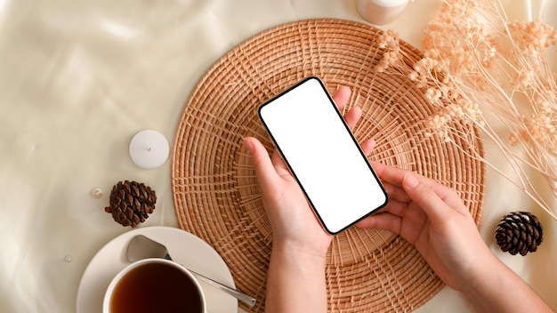 Ansicht von oben Die Hände einer Frau, die ein weißes Smartphone-Bildschirm-Display-Mockup über einem minimalen Arbeitsbereich halten