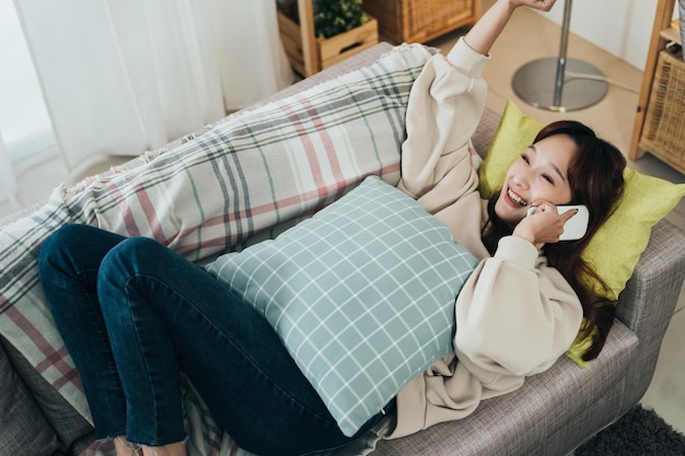 Ansicht von oben Glückliche asiatische Dame, die am Telefon spricht, jubelt mit einem Hebearm über gute Nachrichten, während sie bequem zu Hause auf der Couch im Wohnzimmer liegt.