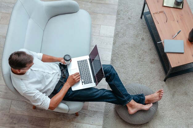 Ansicht von oben. junger Mann mit einer Tasse Kaffee benutzt einen Laptop, während er auf dem Sofa sitzt.