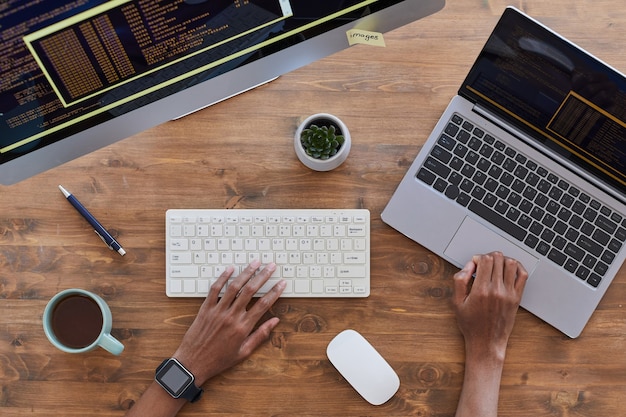 Ansicht von oben nach unten an männlichen Händen, die auf Tastatur am zeitgenössischen Arbeitsplatz mit Computern und Kaffeetasse auf strukturiertem Holztisch tippen, kopieren Raum