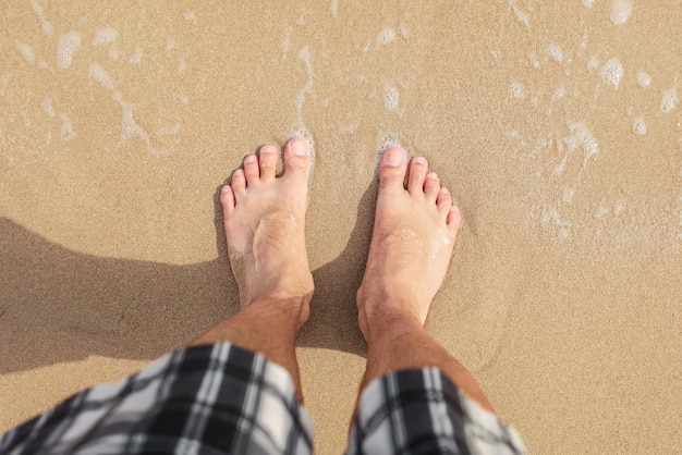 Ansicht von oben nach unten, Wasser wäscht Mannfüße, die am nassen Sand des Strandes stehen, Sonne scheint auf