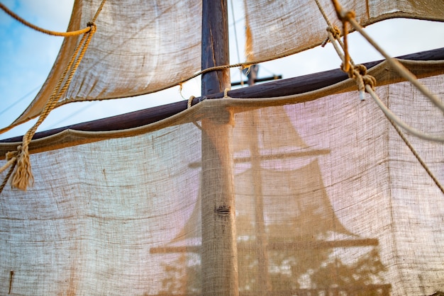 Ansicht von unten auf den Schiffsmast mit beigefarbenen Segeln schwingt gegen den Himmel mit sonnigen, sonnigen, warmen Sommertagwolken