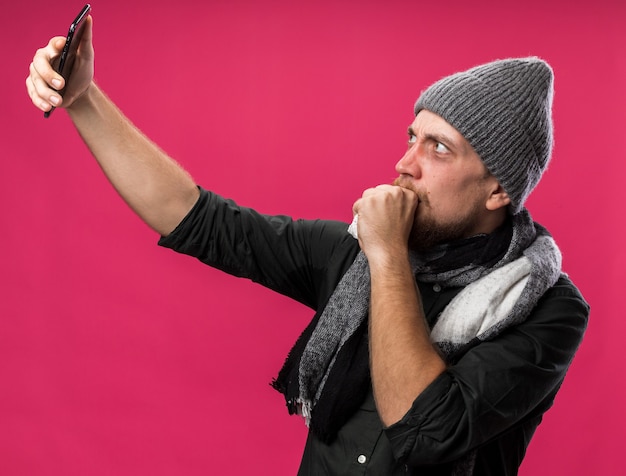 Ansioso joven eslavo enfermo con bufanda alrededor del cuello con sombrero de invierno mantiene el puño cerca de la boca sosteniendo y mirando el teléfono