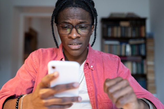 Ansioso y preocupado joven africano sosteniendo un teléfono móvil leyendo sms recibiendo noticias desagradables