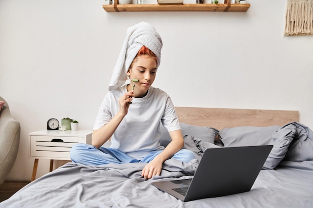 Ansprechende röthaarige Queer-Person in Homeware, die mit einer Gesichtsrolle auf dem Bett mit einem Laptop entspannt