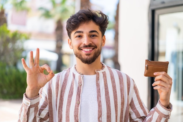 Foto ansprechender arabischer mann hält eine brieftasche im freien und zeigt mit den fingern das ok-zeichen