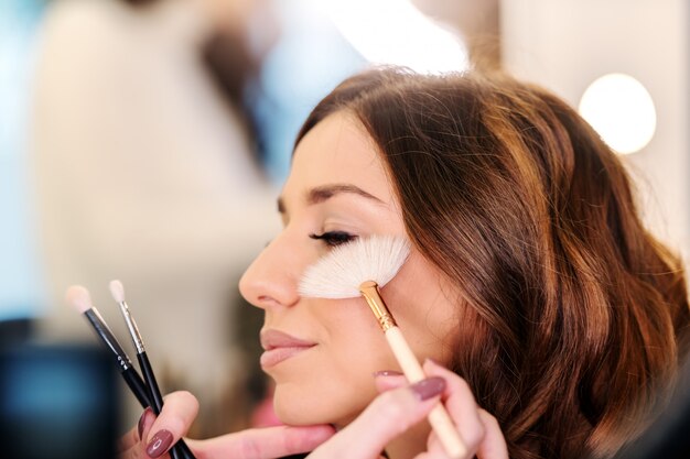 Anspruchsvolle schöne kaukasische Brünette, die im Schönheitssalon sitzt, während Maskenbildner erröten auf Wangen der Frau setzen.