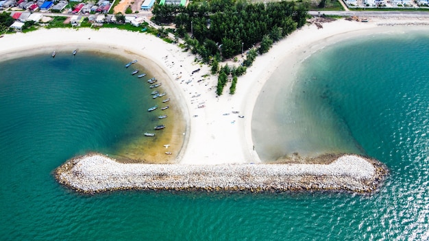 Foto antena de rompeolas de la playa con vista del grupo de personas y pequeño bote de pesca en la playa de arena blanca, árboles y agua de mar clara.