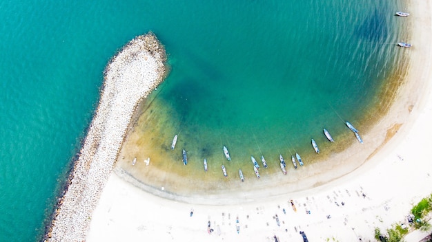 Antena de rompeolas de la playa con vista del grupo de personas y pequeño bote de pesca en la playa de arena blanca, árboles y agua de mar clara.