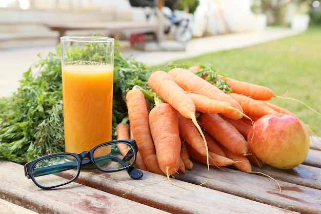 Anteojos y un vaso de jugo de zanahoria y manzana fresco. Frutas y verduras al fondo