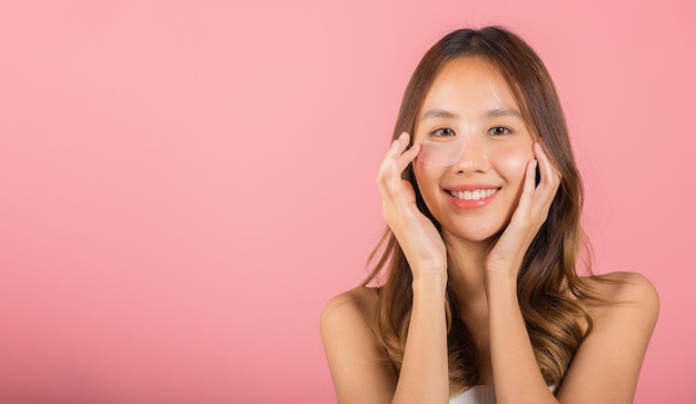 Anti-envelhecimento. Uma jovem asiática bonita e sorridente máscara cosmética gel sob os olhos, Retrato feminino aplicando um tapa-olho de hidrogel médico no rosto, tiro de estúdio isolado em fundo rosa, cuidados com a pele