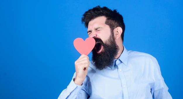 Anti-Valentine-Gähnen Hipster mit kleinem Herzen Schläfriger valentine Mann mit herzförmiger Karte Bärtiger Mann mit rotem Herz Langweiliger Valentinstag Ausdruck seiner Liebe Kopie Raum