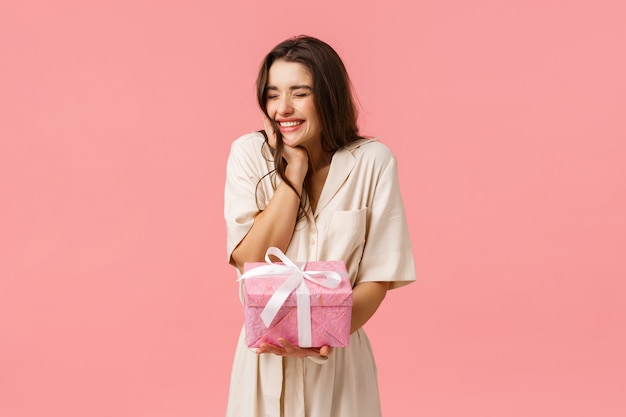 Anticipación, celebración y concepto de fiesta. Alegre hermosa mujer joven en vestido, animando ojos cerrados felizmente sonriendo y riendo, recibiendo un bonito regalo, recibió un bonito regalo, pared rosa