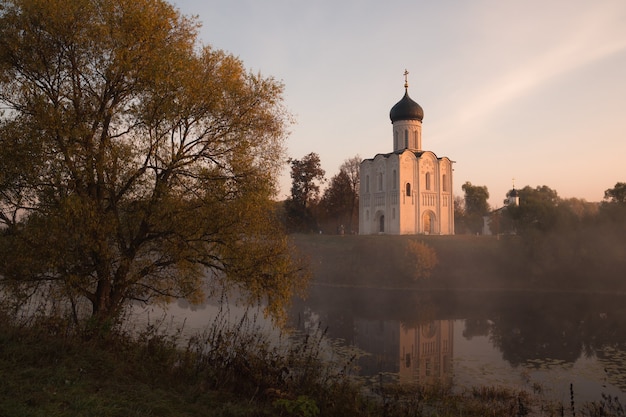 Antiga igreja ortodoxa russa em meio a uma névoa leve ao nascer do sol, refletindo em um lago com uma árvore em primeiro plano