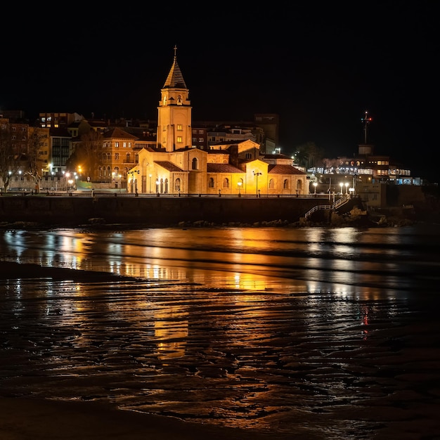 Antigua iglesia de piedra situada junto al mar por la noche con reflejos en el agua Gijón Asturias