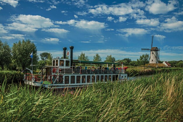 Antiguo barco de vapor y molino de viento en un canal con arbustos cerca de Damme, un encantador pueblo de campo en Bélgica