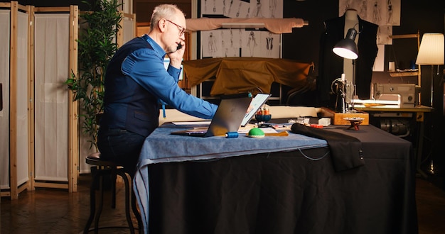 Antiguo diseñador llamando al cliente en el atelier, discutiendo sobre medidas y tela en una llamada telefónica. El sastre senior trabajando en una línea de ropa hecha a medida para crear artículos a medida.