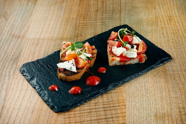 Antipasti italianos tradicionais - bruschetta com queijo feta e tomates em uma placa preta da ardósia em uma superfície de madeira.