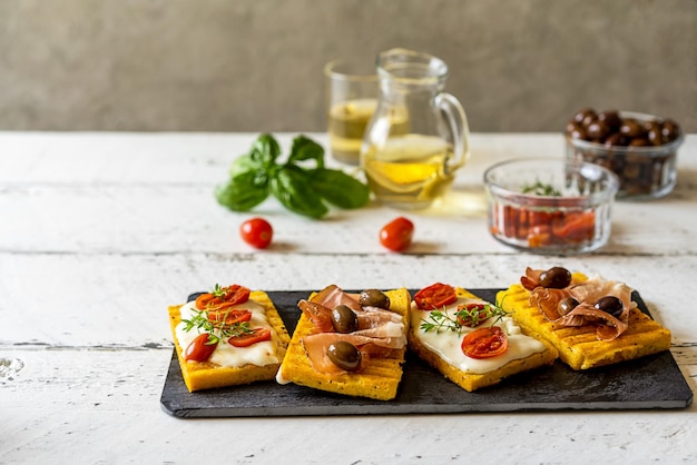 Antipasto con polenta a la parrilla cubierto con queso stracchino, tomates, hierbas, speck y aceitunas