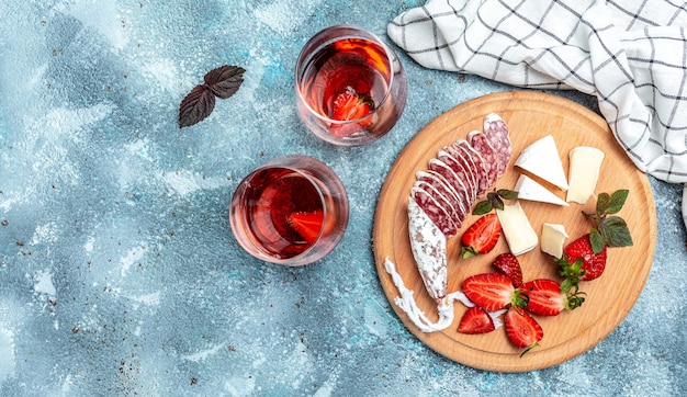 Antipasto en rodajas de salami fuet español wurst, queso camembert, fresas y vino rosado de cristal sobre fondo azul. banner, lugar de receta de menú para texto, vista superior.