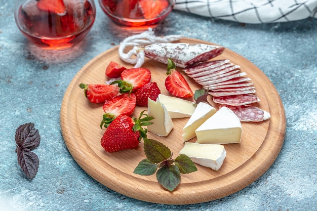 Antipasto en rodajas de salami fuet español wurst, queso camembert, fresas y vino rosado de cristal sobre fondo azul. Fondo de receta de comida. De cerca.