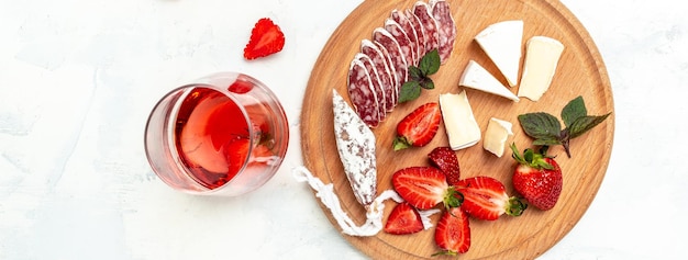 Antipasto en rodajas de salchicha de salami fuet español, queso camembert, fresas y vino rosado de cristal sobre fondo blanco. banner, lugar de receta de menú para texto, vista superior.