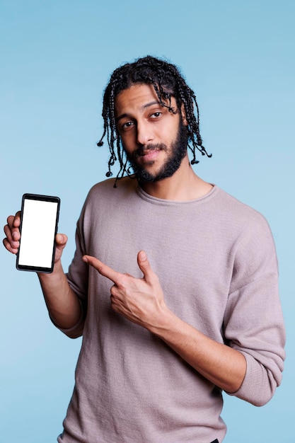 Anunciante árabe seguro que sostiene un teléfono inteligente y apunta a una pantalla táctil blanca vacía mientras mira la cámara. Hombre que presenta un teléfono móvil con pantalla en blanco, producto publicitario