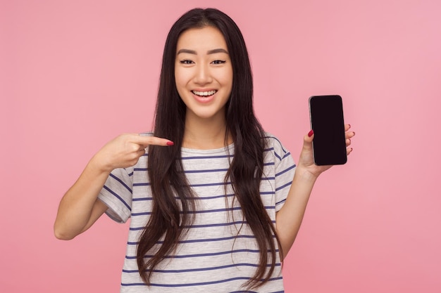 Anuncio de dispositivo móvil o aplicación, Retrato de una chica morena feliz apuntando al teléfono celular y sonriendo a la cámara, satisfecha con el celular. tiro de estudio interior aislado sobre fondo de color rosa