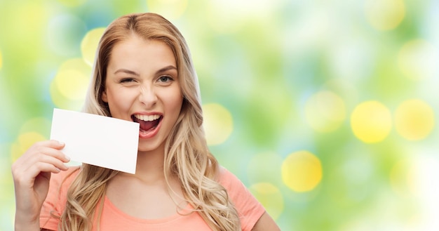 Foto anuncio, invitación, mensaje y concepto de la gente - mujer joven sonriente o adolescente con tarjeta de papel blanco en blanco sobre fondo de luces verdes de verano